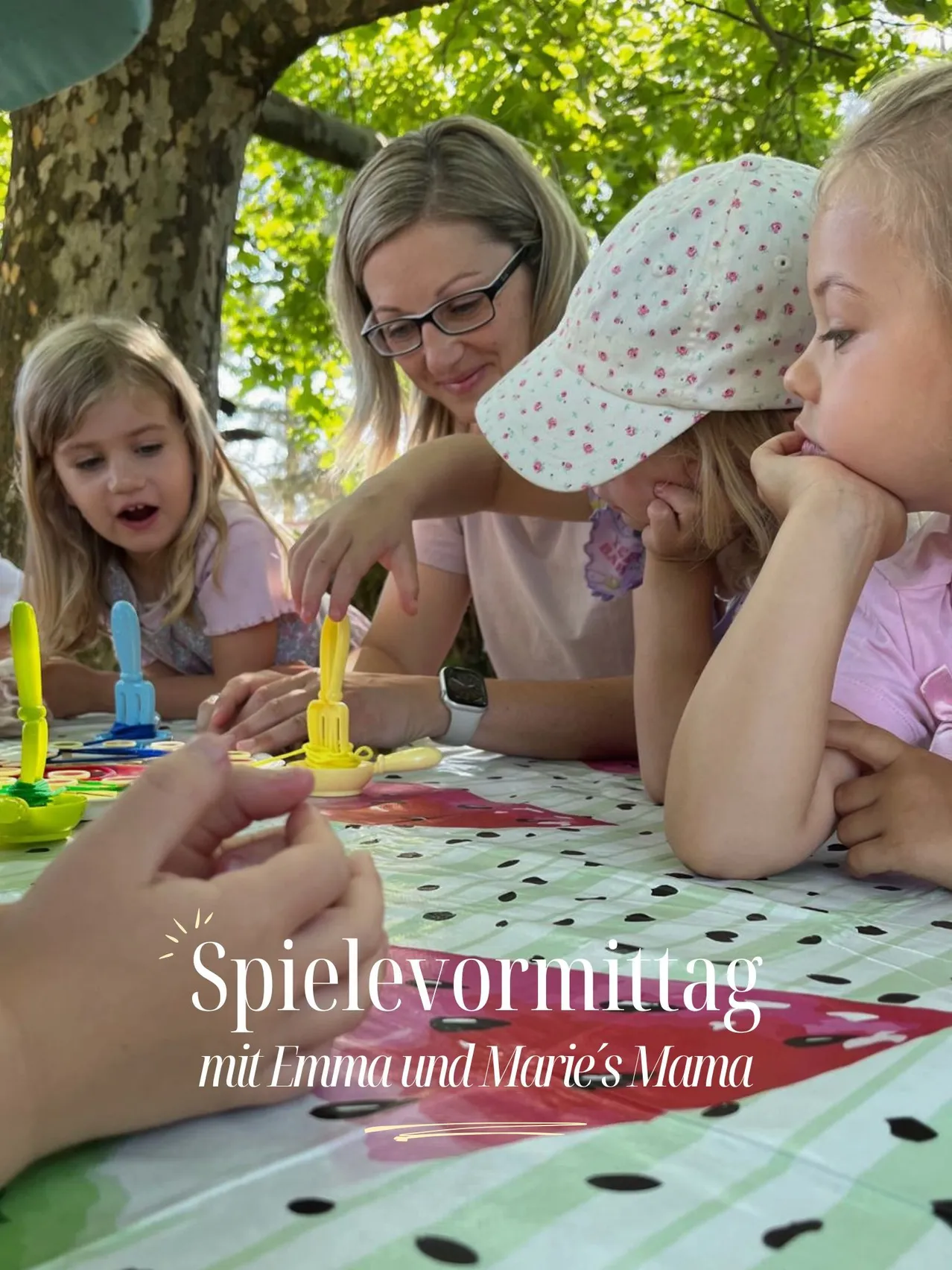 Eine Gruppe von Kindern und Erwachsenen sitzt um einen Tisch mit Spielzeug. Eine Frau mit Brille hält ein Spielzeug, während ein Mädchen mit Mütze zusieht. Die Tischdecke hat ein Wassermelonendesign.