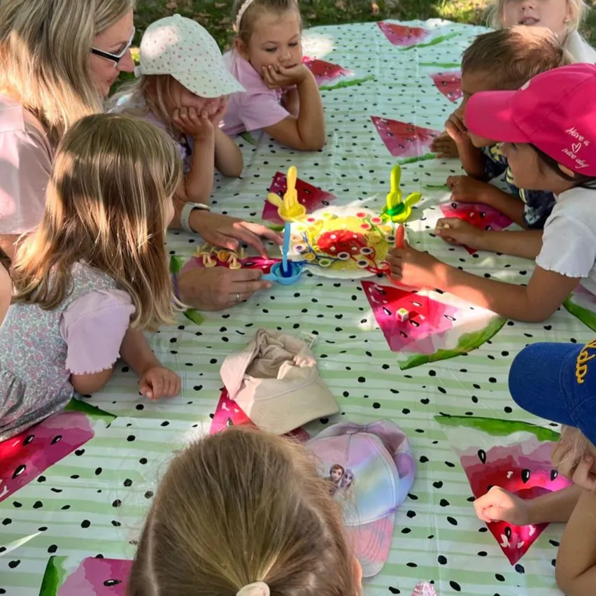 Mehrere junge Kinder und eine Frau versammeln sich um einen Tisch, der mit einem Tupfentuch mit Wassermelonenmuster bedeckt ist. Die Kinder beschäftigen sich mit Spielzeug und Bastelmaterialien.