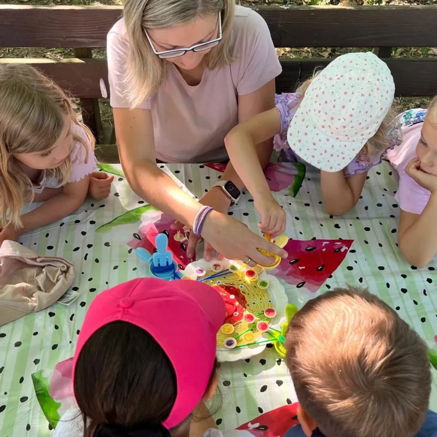 Eine Frau und mehrere Kinder sitzen um einen Tisch mit einem gepunkteten Tischtuch und genießen eine Mahlzeit. Die Frau trägt eine Brille und ein Armband, während die Kinder Hüte und Mützen tragen.