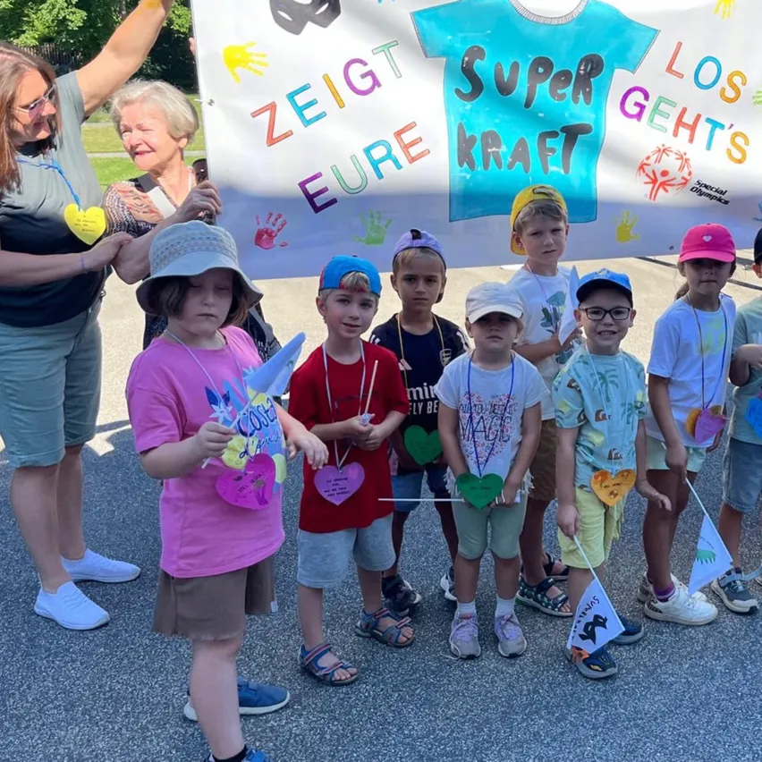 Eine Gruppe von Kindern und Erwachsenen steht vor einem Banner mit bunten Handabdrücken. Das Banner sagt 'Zeig deine Kraft' auf Deutsch. Sie halten Fahnen und tragen Medaillen.