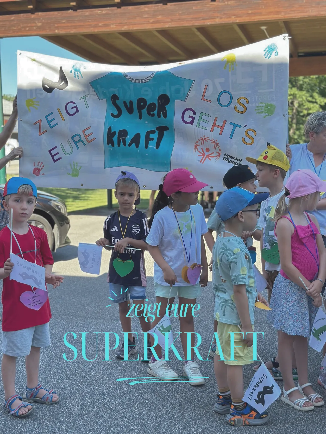 Eine Gruppe von Kindern steht mit Bannern und Schildern mit den Worten 'Superkraft'. Sie tragen bunte Mützen und Uniformen. Der Hintergrund zeigt ein Banner mit demselben Text.