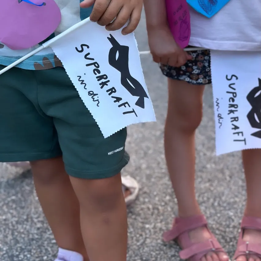 Ein Kind hält eine weiße Fahne mit einer schwarzen Maskensilouette. Ein weiteres Kind dahinter hält eine ähnliche Flagge. Beide tragen Blumenshorts und Sandalen.
