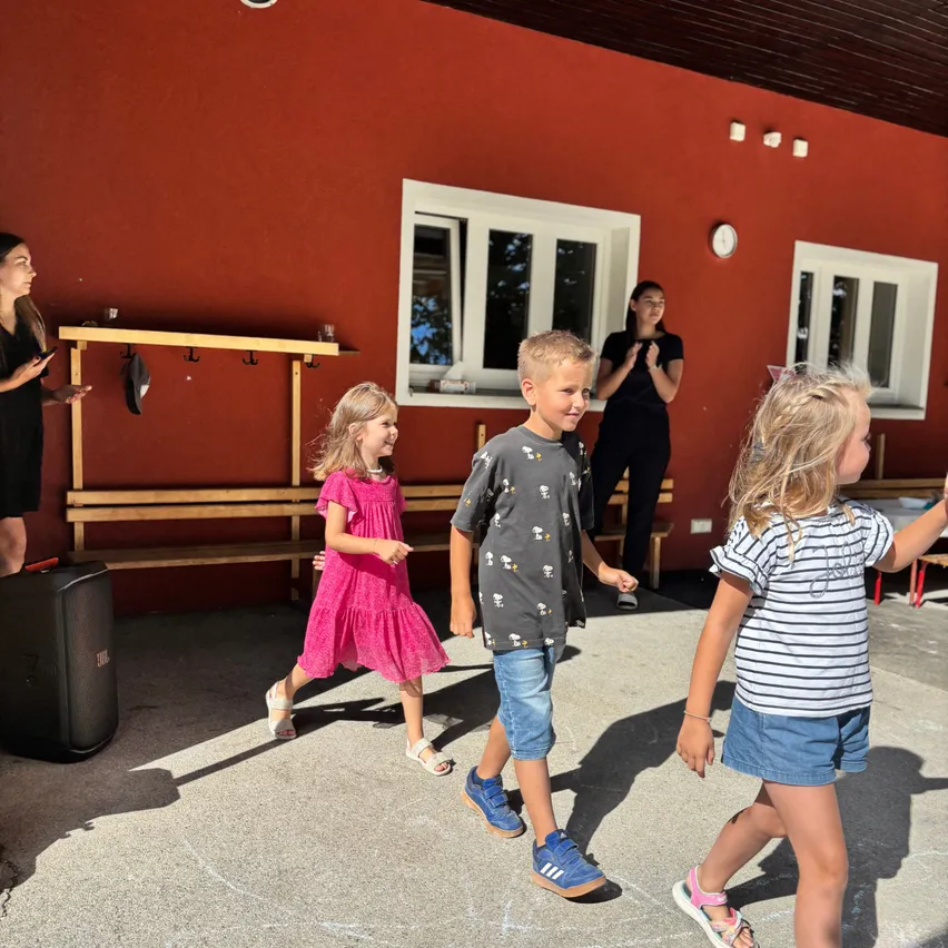 Drei Kinder gehen in einem Innenhof mit einer roten Wand, einer Holzbank und einer Frau, die in der Nähe einer Uhr steht.