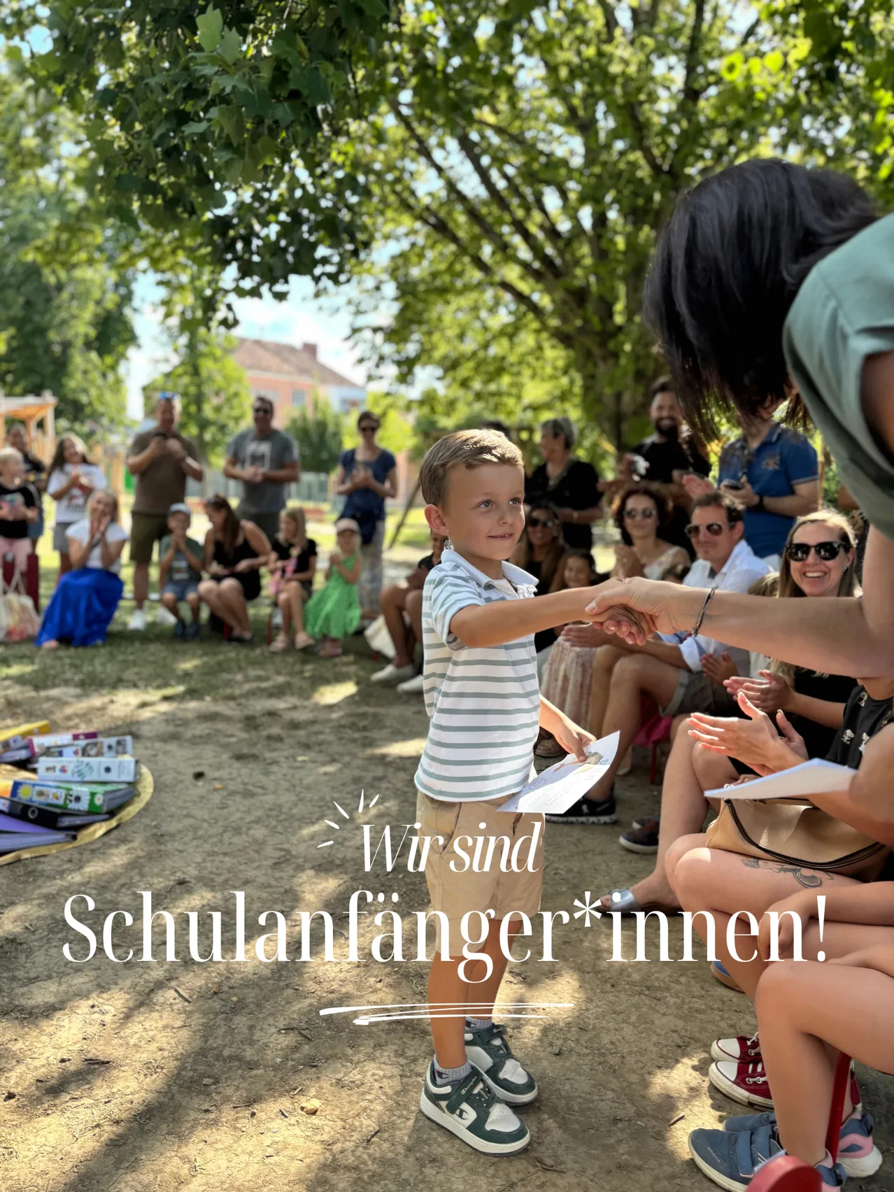 Ein junger Junge erhält ein Zertifikat in einem Park, umgeben von einer Menschenmenge, einige sitzend und andere stehend, unter einem Baum.