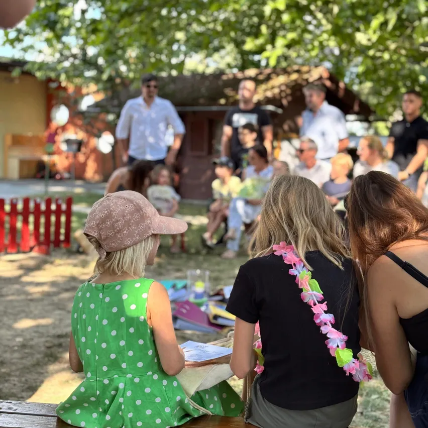 Eine Gruppe von Menschen sitzt auf einer Bank unter einem Baum. Ein Mädchen in einem grünen Punktekleid sitzt vorne, und ein Mädchen in einem schwarzen Shirt mit einer Blumengirlande sitzt neben ihr.