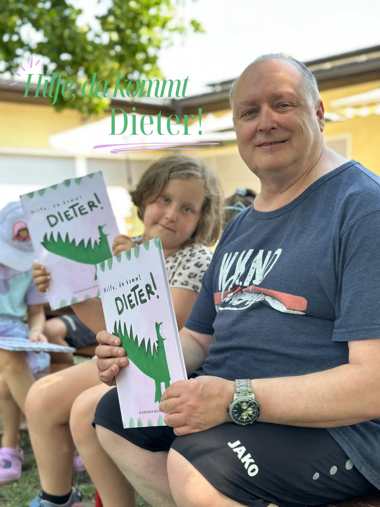 Ein Mann lächelt, während er ein Buch mit dem Titel 'Hilfe, da kommt Dieter!' mit einem Dinosaurier auf dem Cover hält, neben einem kleinen Mädchen, das ein ähnliches Buch in der Hand hat. Im Hintergrund ist verschwommen ein Gebäude und ein Baum zu sehen.