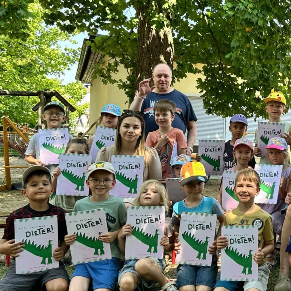 Eine Gruppe von Kindern und Erwachsenen posiert für ein Foto in einem Park und hält Schilder mit der Aufschrift 'Dieter!' und Dinosaurier-Illustrationen hoch.