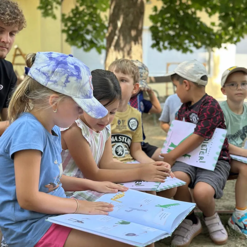 Eine Gruppe von Kindern sitzt auf einer Bank unter einem Baum und liest Bücher. Ein Mädchen in einem blauen Shirt und einer Kappe hält ein Buch mit einem Dinosaurier auf dem Cover.