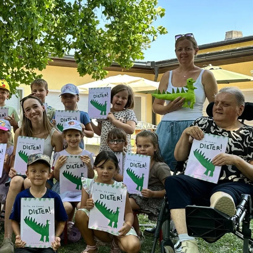 Eine Gruppe von Kindern und Erwachsenen posiert für ein Foto mit Büchern und einem grünen Dinosaurier-Spielzeug. Die Bücher haben das Wort 'Dieter' darauf.