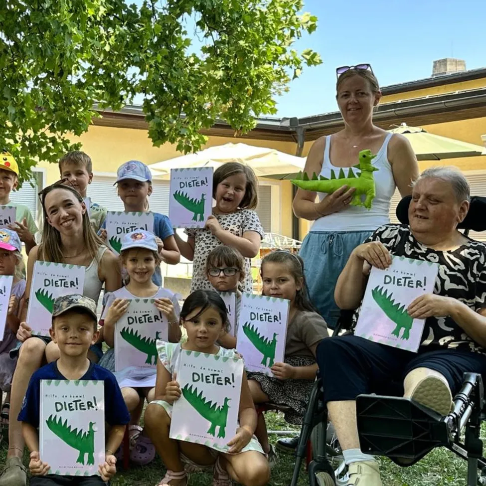 Eine Gruppe von Kindern und Erwachsenen posiert für ein Foto mit Büchern und einem grünen Dinosaurier-Spielzeug. Die Bücher haben das Wort 'Dieter' darauf.