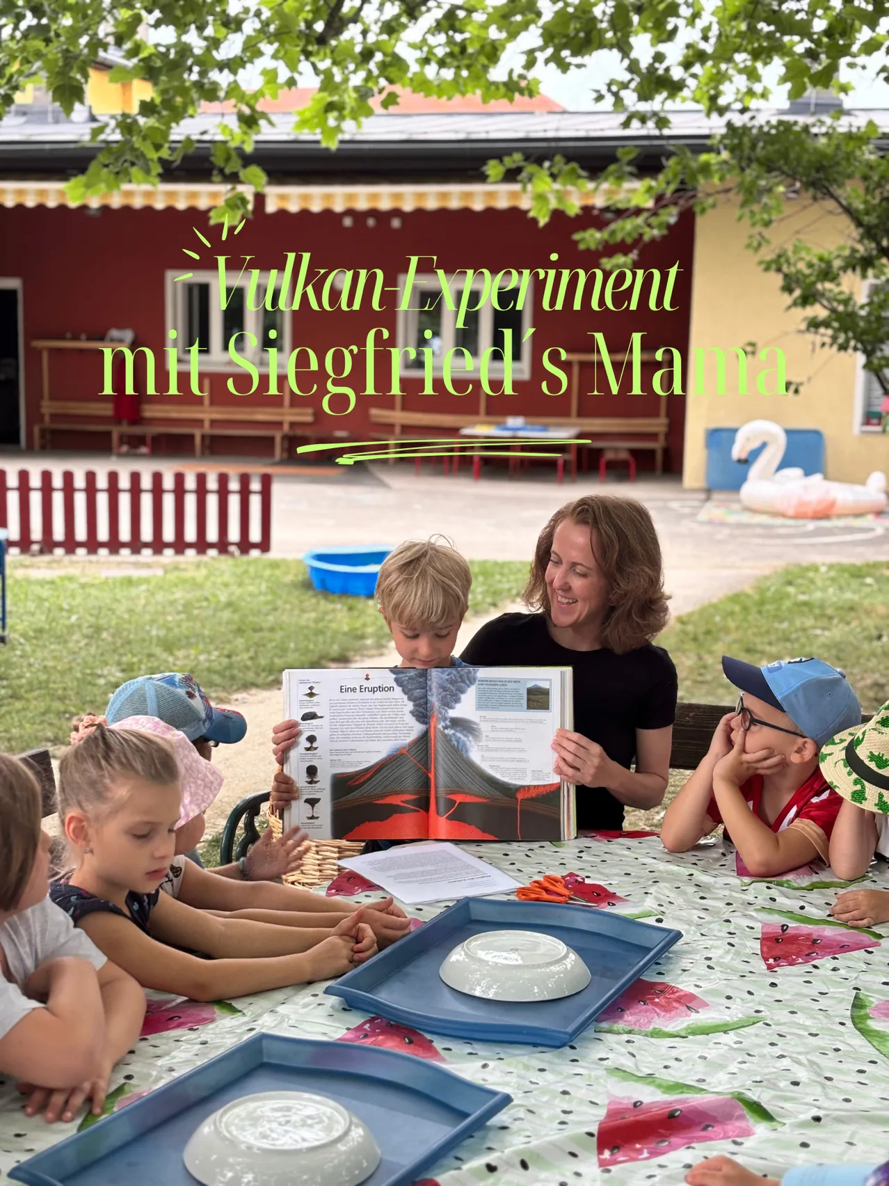 Eine Frau liest ein Buch über Vulkane für eine Gruppe von Kindern, die um einen Tisch im Freien sitzen. Der Tisch hat ein blaues Tablett und eine Schüssel. Der Textüberlagerung lautet 'Vulkan Experiment mit Siegfried's Mama.'