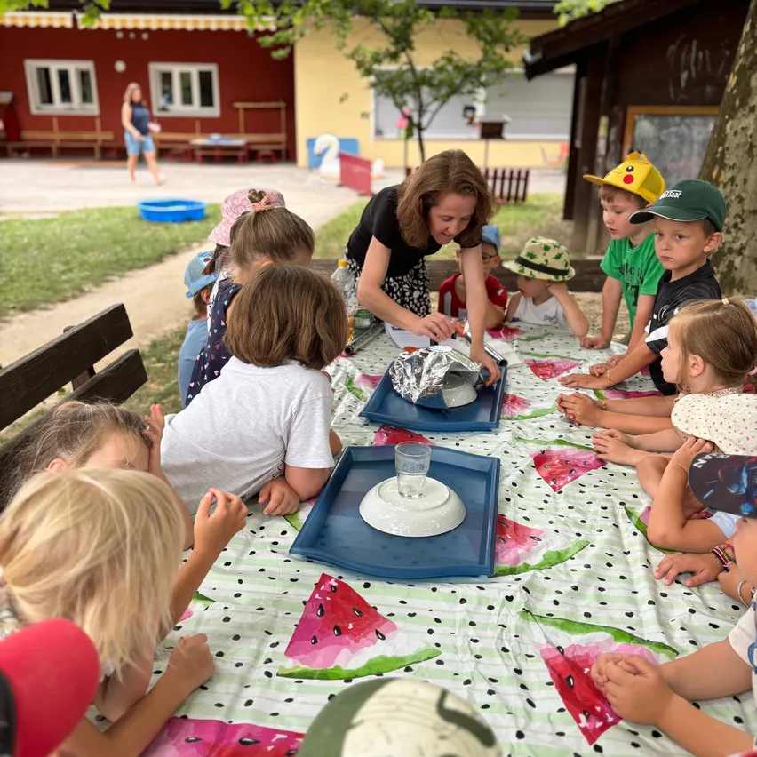 Eine Gruppe von Kindern sitzt um einen Tisch mit einem Wassermelonen-Thema, während eine Frau Essen auf einem blauen Tablett zubereitet.