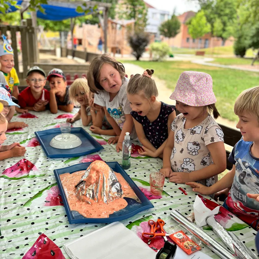 Kinder sitzen um einen Tisch mit einem Vulkanmodell und beobachten gespannt, wie es ausbricht. Sie sitzen in einem Park mit Bäumen und Gebäuden im Hintergrund.