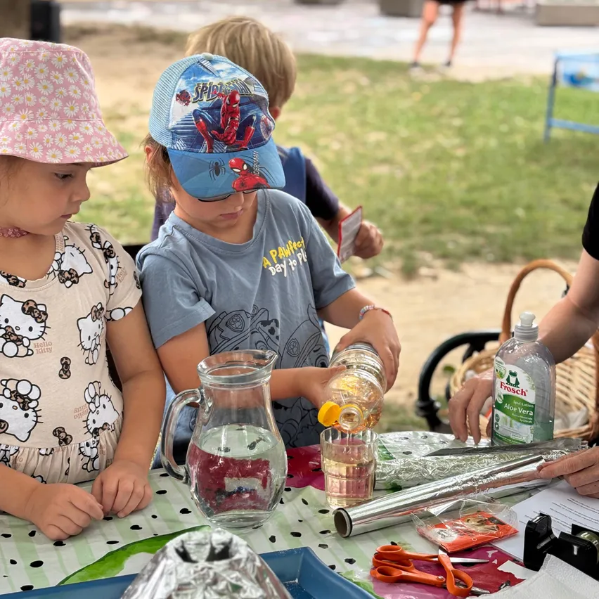 Zwei Kinder sitzen an einem Tisch, eines gießt eine Flüssigkeit in ein Glas, während das andere zuschaut. Verschiedene Gegenstände, einschließlich einer Karaffe und Scheren, sind auf dem Tisch. Eine Person im Hintergrund ist teilweise sichtbar.
