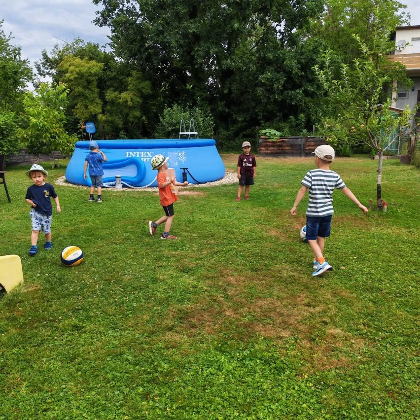 Kinder spielen Fußball im Garten mit einem aufblasbaren Pool im Hintergrund.