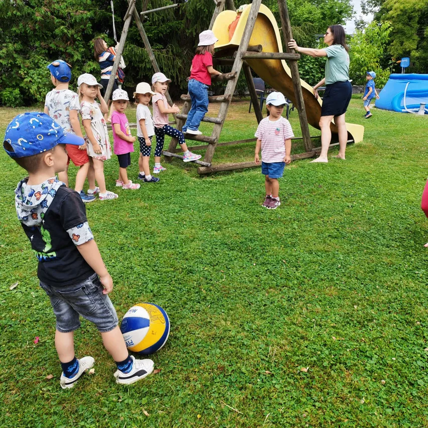 Eine Gruppe von Kindern spielt im Garten mit einer Rutsche und einem Ball. Eine Frau steht in der Nähe.