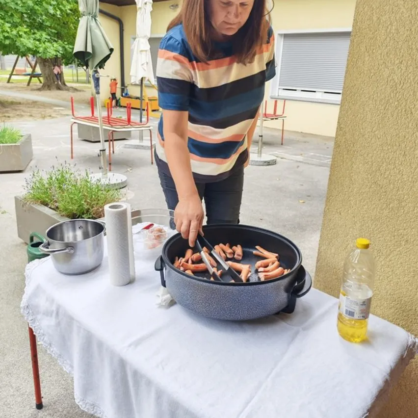 Eine Frau in einem gestreiften Shirt kocht Würstchen in einem Topf auf einem Tisch draußen.