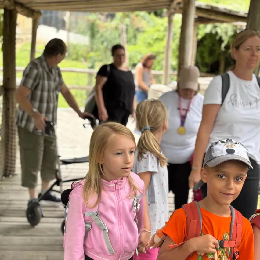 Eine Gruppe von Menschen, darunter Kinder, geht auf einem Holzweg. Die Kinder tragen Rucksäcke und scheinen auf einer Schulfahrt zu sein. Die Erwachsenen stehen hinter ihnen.