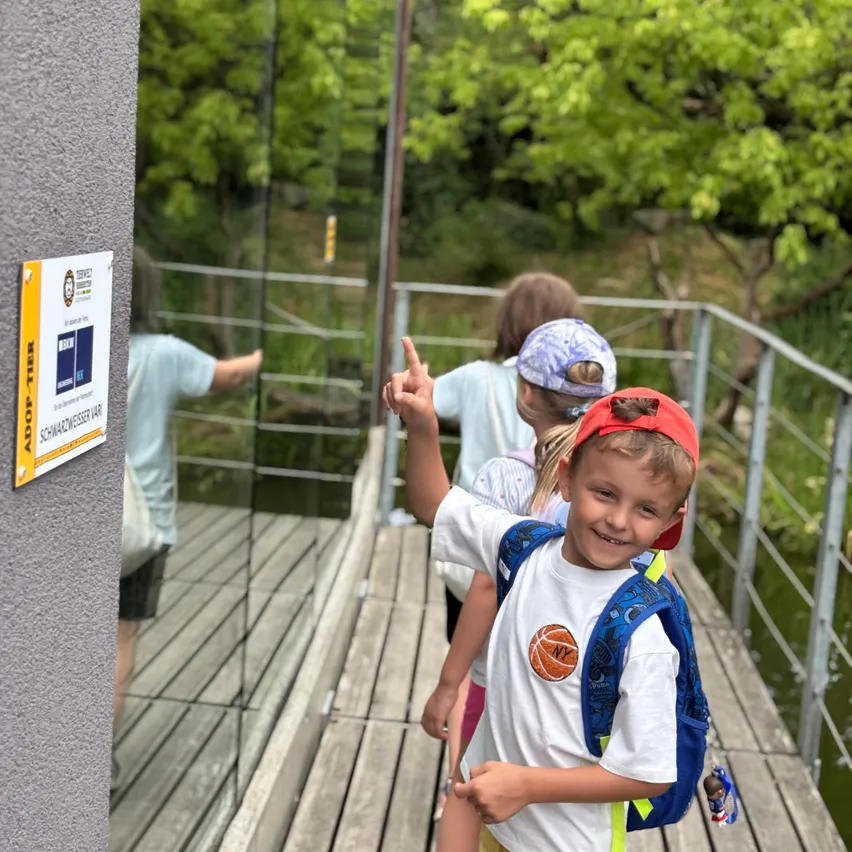 Ein Junge mit Rucksack und rotem Hut lächelt und zeigt auf ein Schild an einer Wand. Hinter ihm gehen ein Mädchen mit blauer Mütze und ein weiterer Junge über eine Holzbrücke.