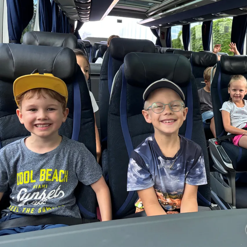 Zwei Jungen sitzen nebeneinander in einem Bus, lächeln und posieren für ein Foto. Einer trägt eine gelbe Kappe und ein graues T-Shirt mit 'Cool Beach', der andere eine blaue Kappe und ein blaues T-Shirt mit einem Himmelsdesign.
