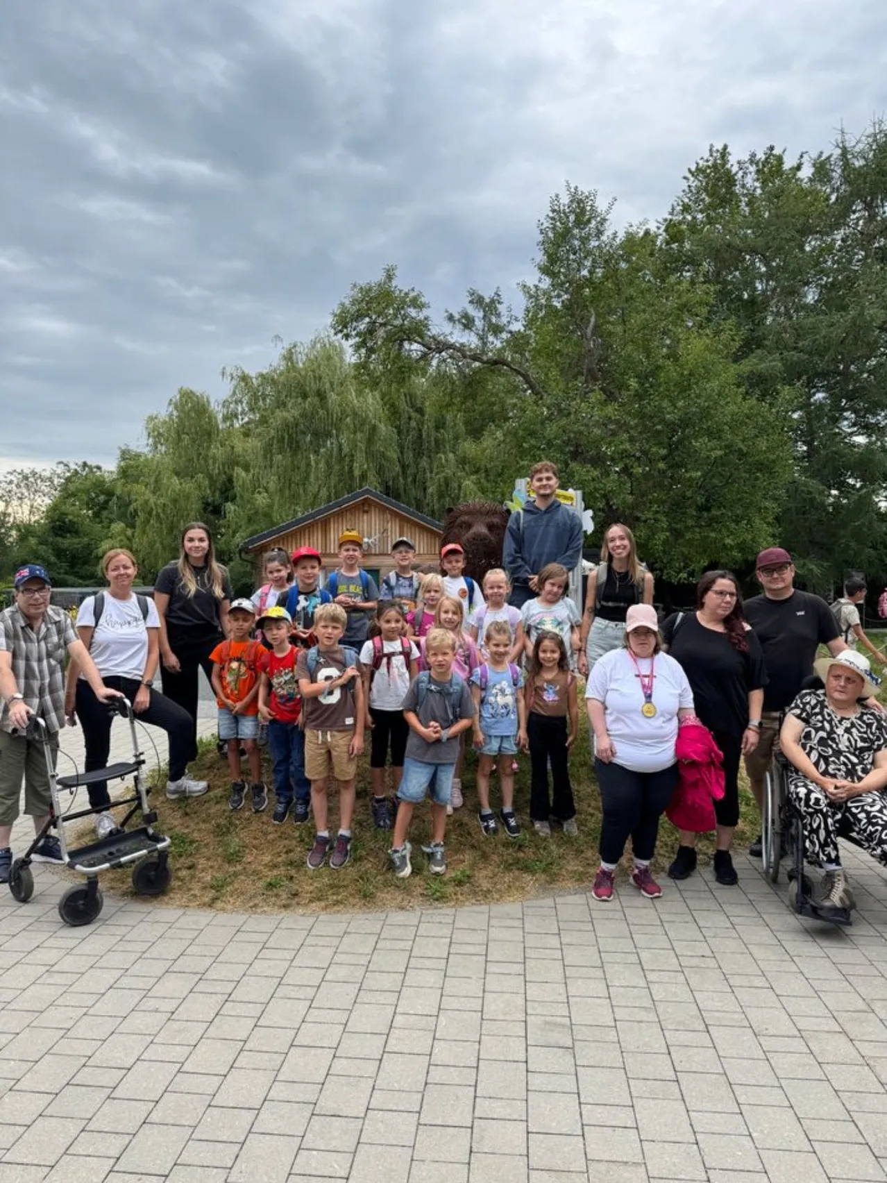 Eine Gruppe von Kindern und Erwachsenen posiert für ein Foto vor einem kleinen Haus mit einer Bärenstatue, unter einem bewölkten Himmel.
