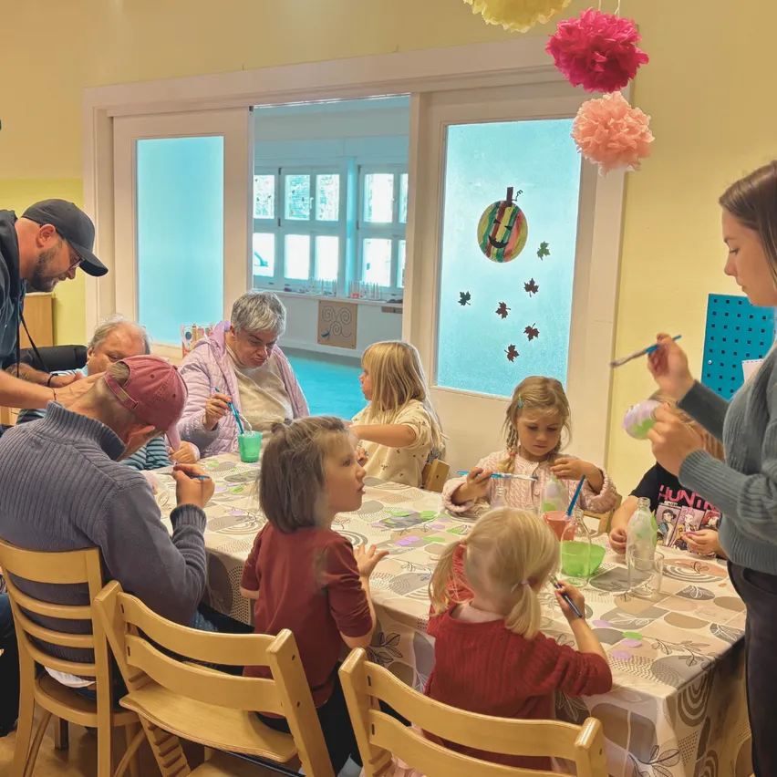 Eine Gruppe von Kindern und Erwachsenen versammelt sich um einen Tisch und beschäftigt sich mit einer Kunstaktivität. Der Tisch ist mit einer gemusterten Tischdecke bedeckt und die Kinder benutzen Pinsel und Becher. Der Raum hat gelbe Wände und dekorative Papierblumen hängen von der Decke.