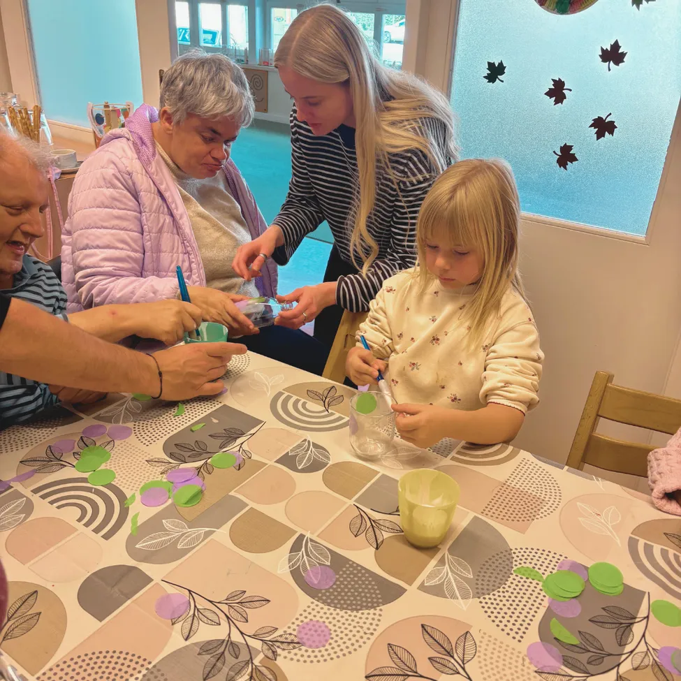 A group of people, including an elderly woman and a young girl, are gathered around a table, engaged in a craft activity. They are holding various art supplies and appear to be creating something.