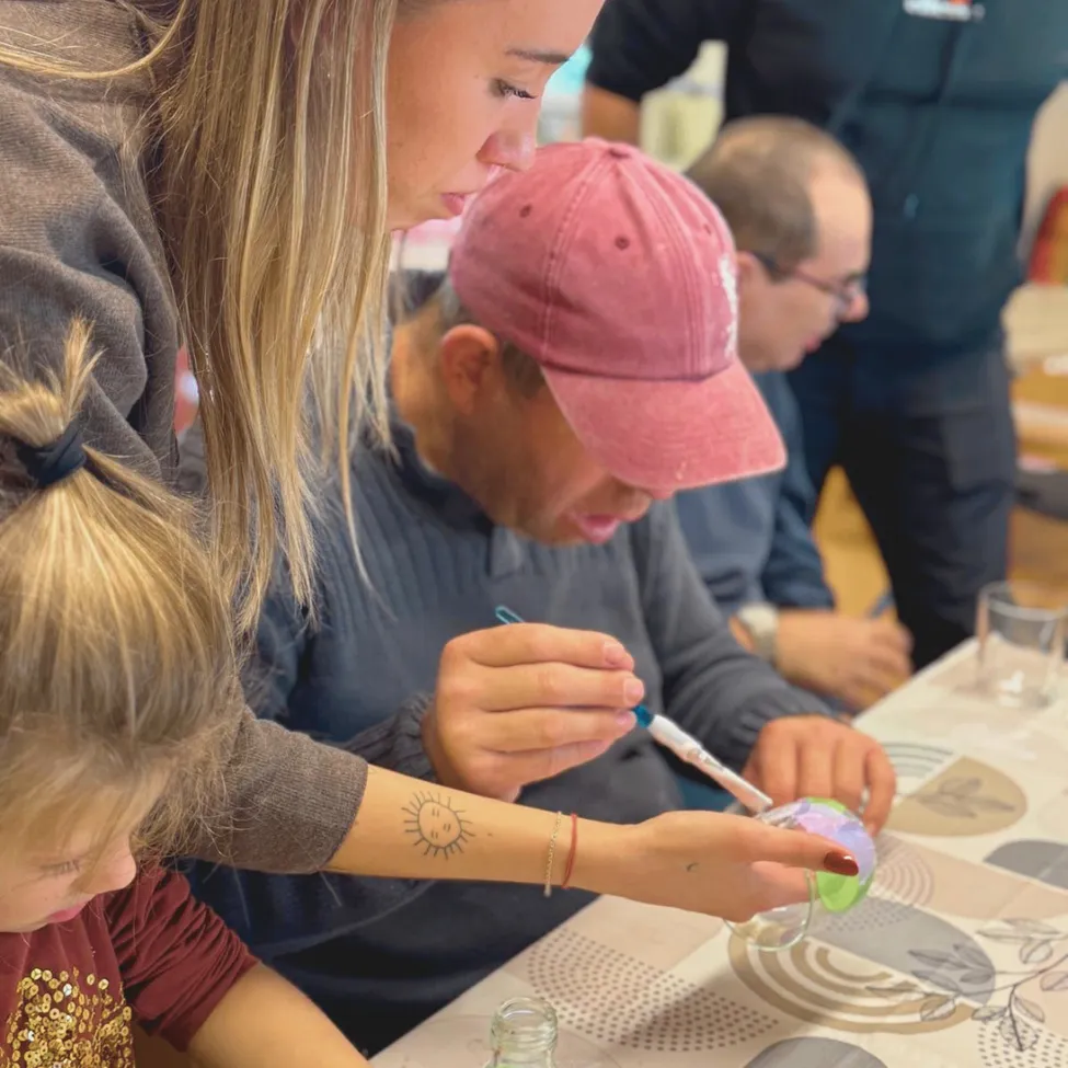 A group of people are gathered around a table, engaged in a craft activity. A woman is helping a young girl with a craft project.