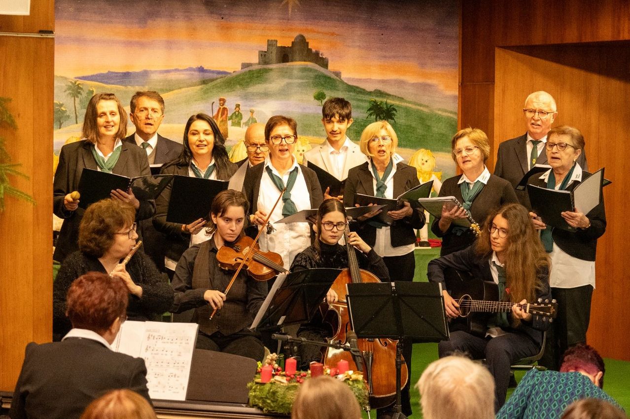 Ein Chor aus Erwachsenen und Kindern tritt auf einer Bühne vor einem Publikum auf, spielt Violine, Cello und Gitarre, mit Notenständern und einem festlichen Kranz im Vordergrund.