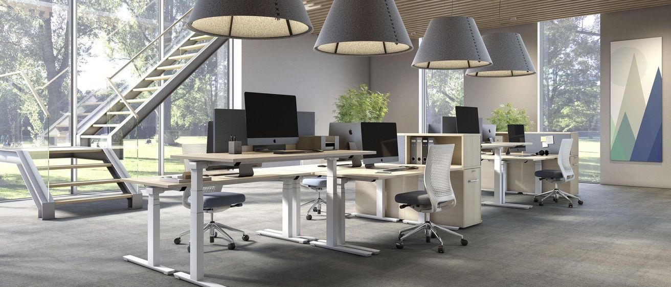 Ein Büroraum mit Stehschreibtischen, grauen Stühlen und großen Fenstern. Mehrere Computer und Monitore stehen auf den Schreibtischen. Der Raum hat Holzdecken und Pendelleuchten.