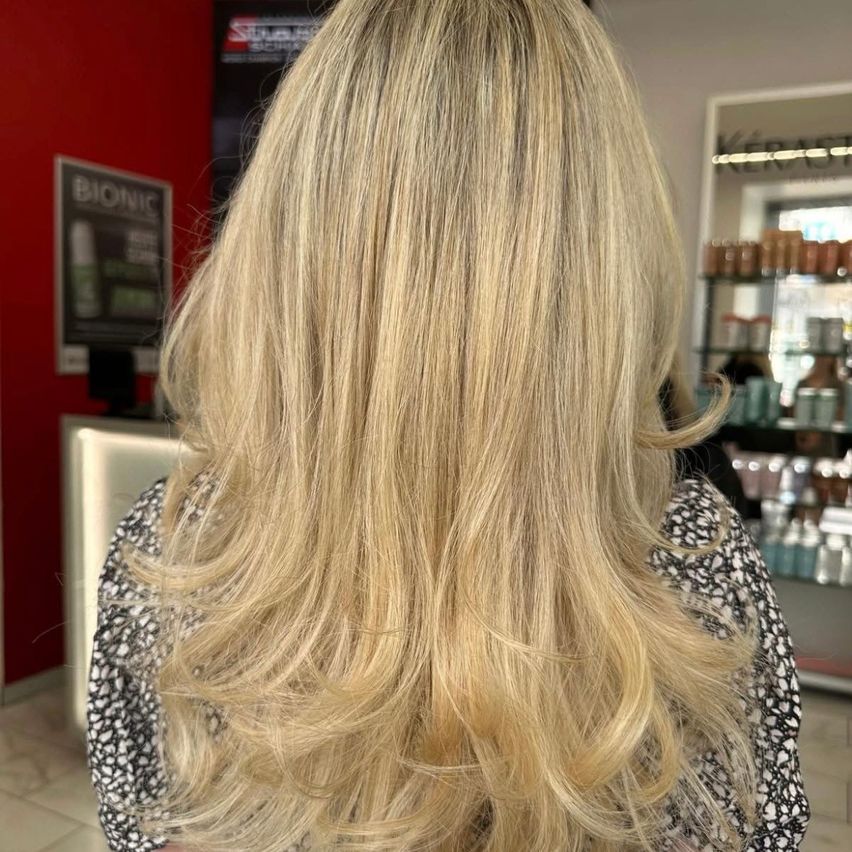 Eine Frau mit langen, hellblonden Haaren, die in Wellen gestylt sind, wird von hinten in einem Salon gezeigt. Dahinter befindet sich eine rote Wand mit einem Schild für Bionic.