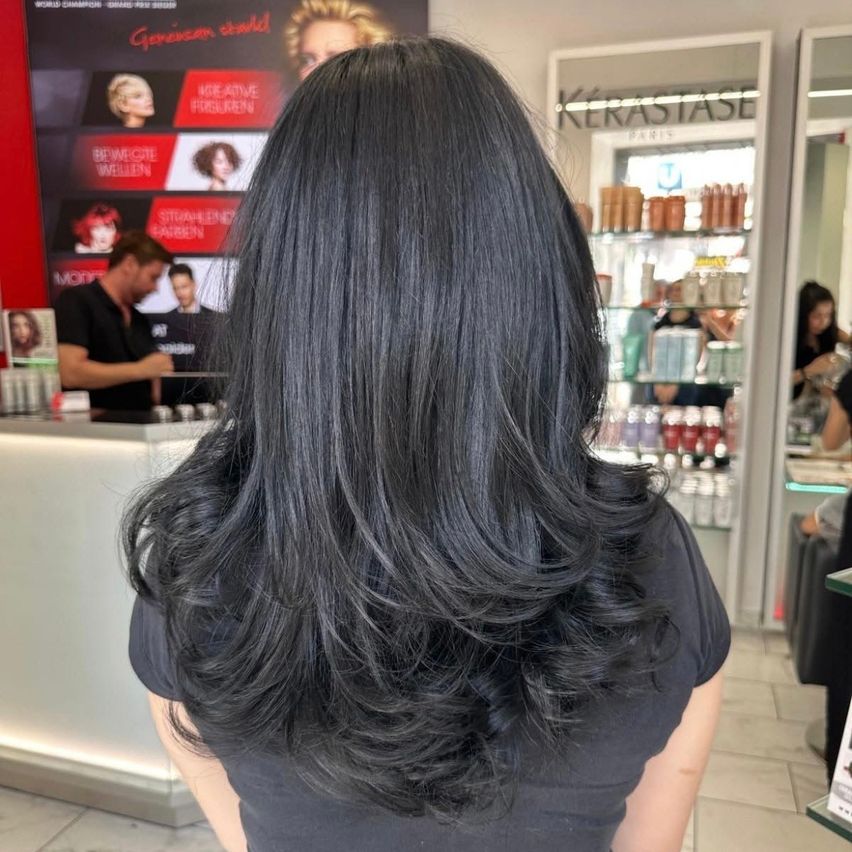 Eine Frau mit langen, welligen schwarzen Haaren steht in einem Salon, mit einem Kérastase-Schild im Hintergrund und einem Mann hinter einem Tresen.