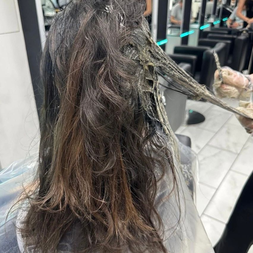 Eine Frau mit langen braunen Haaren lässt sich im Salon die Haare waschen. Die Haare sind nass, und eine Person wäscht sie. Dahinter befinden sich Spiegel und Stühle.