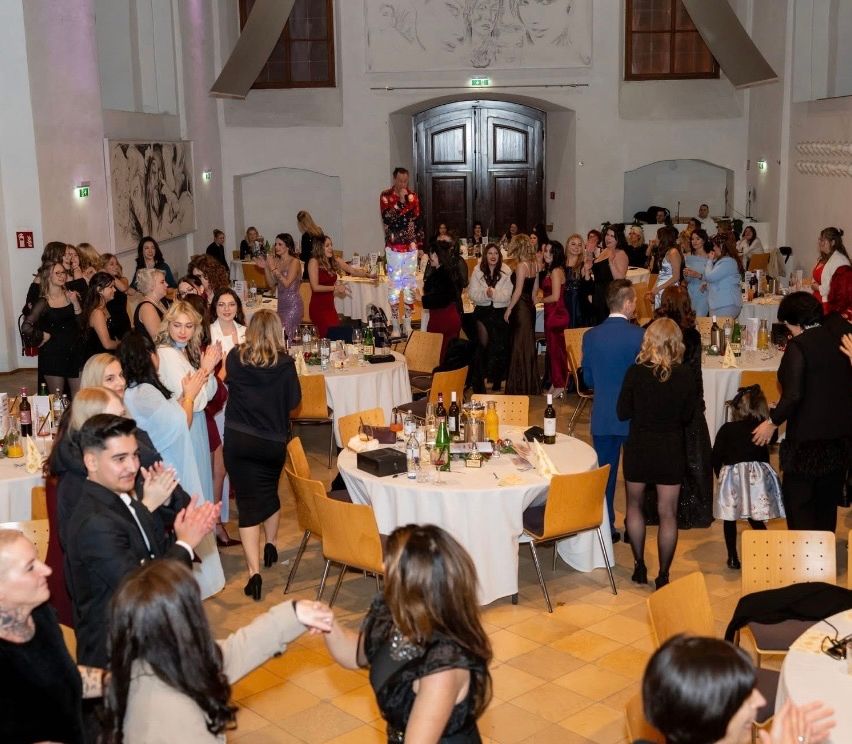 Eine große Gruppe von Menschen bei einer Indoor-Veranstaltung, einige klatschen. Ein Mann steht auf einem Stuhl und trägt ein farbenfrohes Outfit. Der Raum hat runde Tische mit Flaschen und anderen Gegenständen.