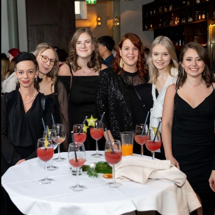 Eine Gruppe von Frauen in schwarzen Kleidern lächelt für die Kamera bei einer Veranstaltung, mit Getränken und Dekorationen auf einem Tisch vor ihnen.