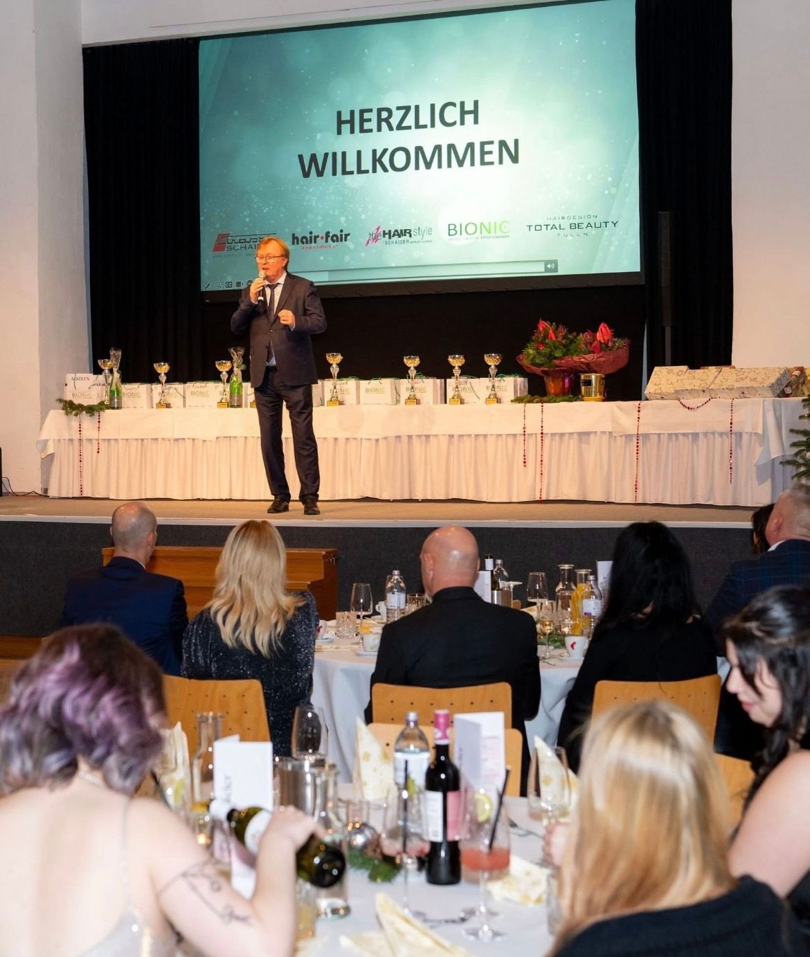 Ein Mann steht auf der Bühne und hält eine Rede, mit einer Leinwand im Hintergrund. Trophäen sind auf der Bühne. Viele Menschen sitzen an Tischen, einige trinken.