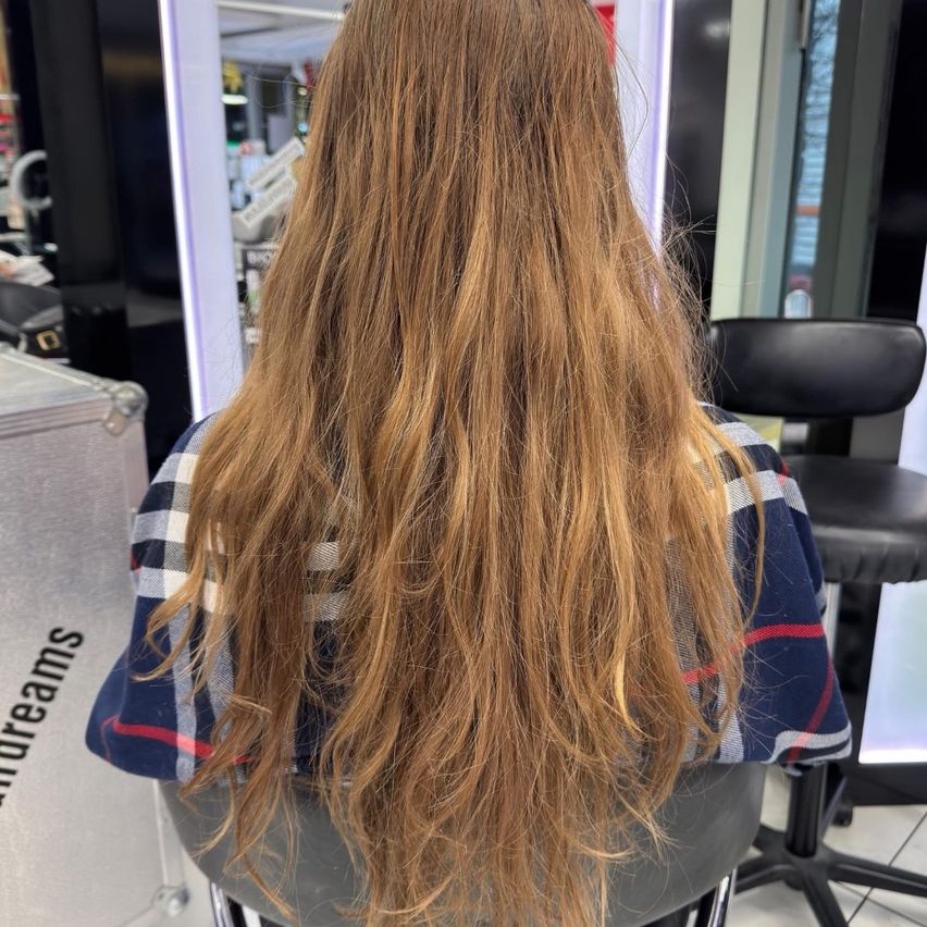 Eine Frau mit langen, welligen blonden Haaren sitzt in einem Salonstuhl, dem Spiegel gegenüber. Hinter ihr befindet sich ein schwarzer Stuhl und eine Glastür.