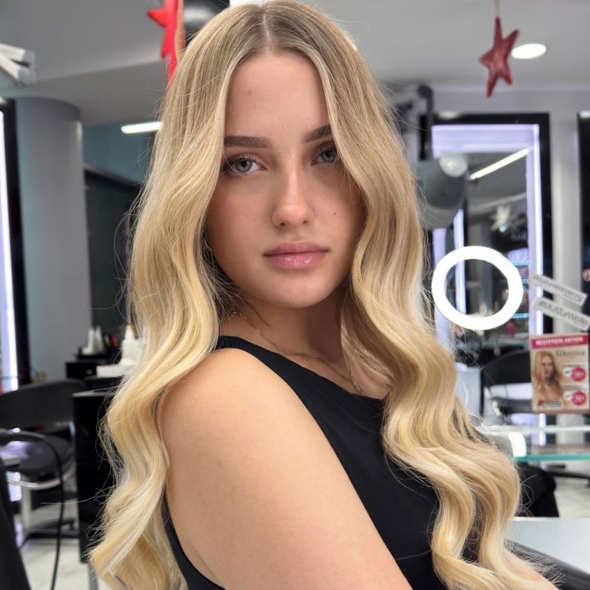 Eine Frau mit langen, blonden, welligen Haaren posiert in einem Salon und trägt ein schwarzes ärmelloses Oberteil. Hinter ihr befinden sich Spiegel, Stühle und eine Produktanzeige.