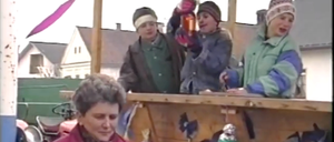 Vier Personen sitzen auf einer Holzplattform, eine hält einen Becher und eine andere ein Glas. Eine Frau in einem roten Mantel schaut direkt in die Kamera. Hinter ihnen sind Häuser zu sehen.