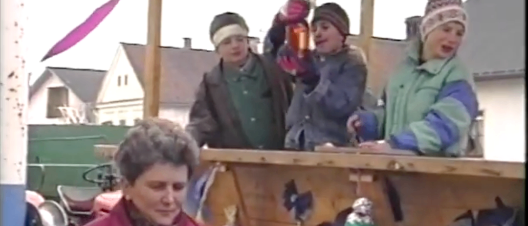 Vier Personen sitzen auf einer Holzplattform, eine hält einen Becher und eine andere ein Glas. Eine Frau in einem roten Mantel schaut direkt in die Kamera. Hinter ihnen sind Häuser zu sehen.