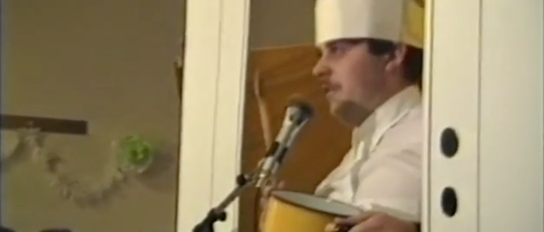 Ein Mann in einer Kochmütze steht vor einem Mikrofon und hält einen gelben Topf. Dahinter ist ein verschwommenes Bild eines anderen Mannes und eines Ventilators zu sehen. Am unteren Rand ist das Datum '4 1 199' angezeigt.