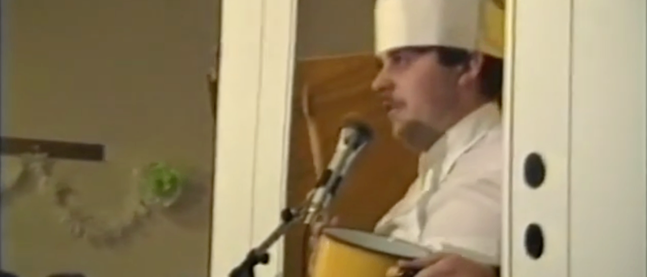 Ein Mann in einer Kochmütze steht vor einem Mikrofon und hält einen gelben Topf. Dahinter ist ein verschwommenes Bild eines anderen Mannes und eines Ventilators zu sehen. Am unteren Rand ist das Datum '4 1 199' angezeigt.