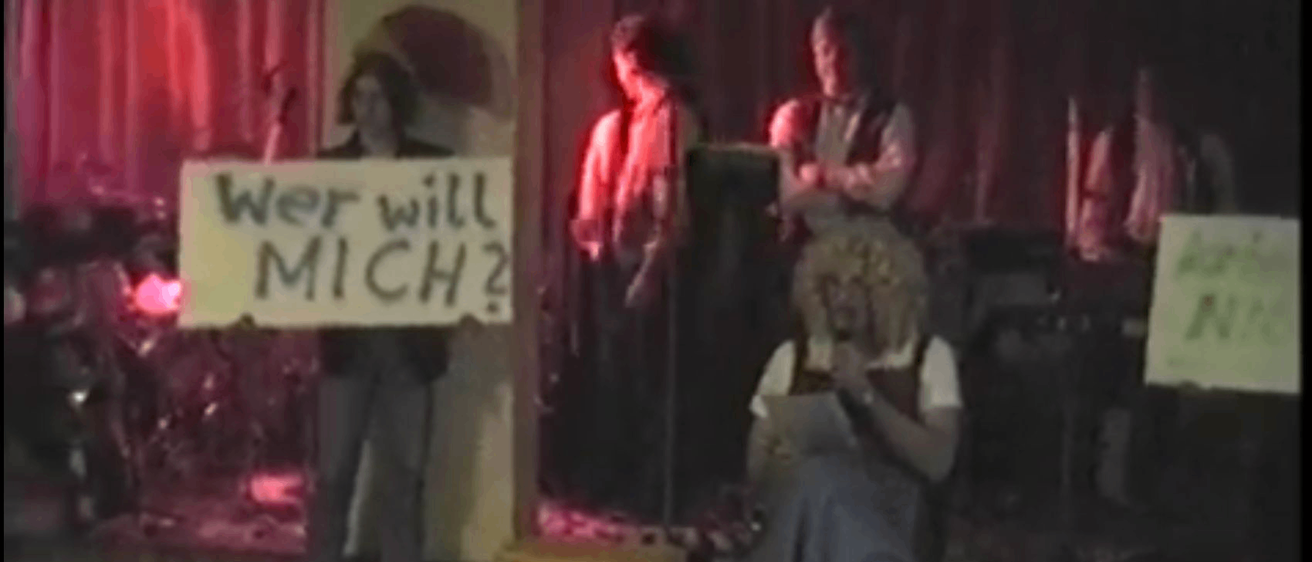 Eine Person hält ein Schild mit der Aufschrift 'Wer will MICH?' Eine Frau sitzt und liest ein Buch, während andere hinter ihr stehen. Ein Datum von 7.1.1996 ist angezeigt.