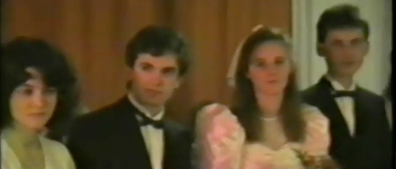 Ein Mann und eine Frau stehen eng beieinander. Der Mann trägt einen schwarzen Anzug und eine Fliege. Die Frau trägt ein rosa Kleid und einen Schleier.