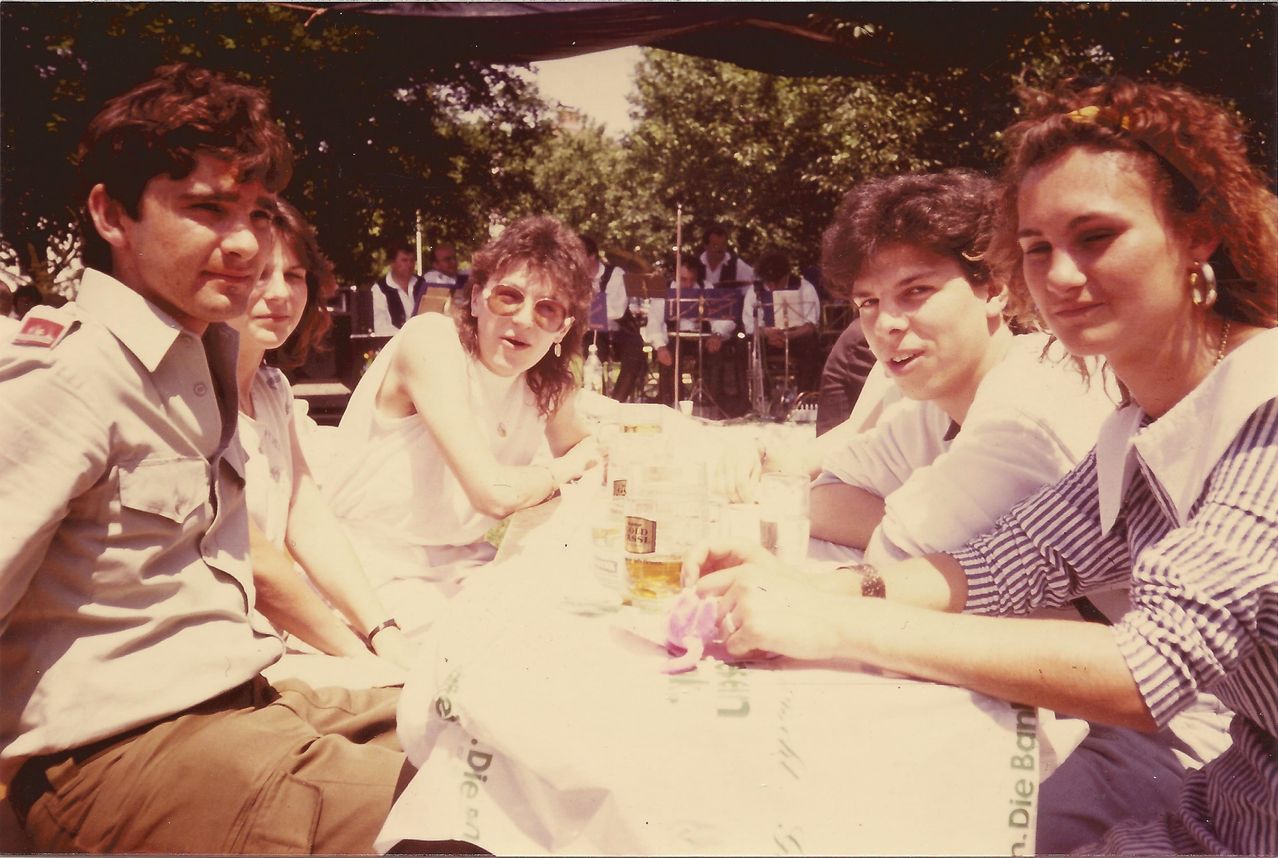 Eine Gruppe von Menschen sitzt draußen an einem Tisch. Eine Frau mit einer Brille und einem weißen Shirt ist in der Mitte. Der Tisch hat Biergläser und Tassen. Hinter ihnen spielen Musiker Instrumente.