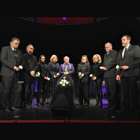Eine Gruppe von Menschen in Schwarz steht auf der Bühne und hält Blumen. Ein Sarg steht in der Mitte, und sie sind wahrscheinlich auf einer Beerdigung.