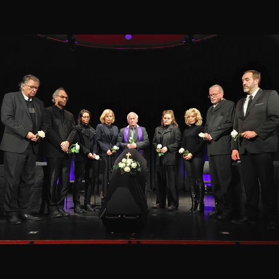 Eine Gruppe von Menschen in Schwarz steht auf der Bühne und hält Blumen. Ein Sarg steht in der Mitte, und sie sind wahrscheinlich auf einer Beerdigung.
