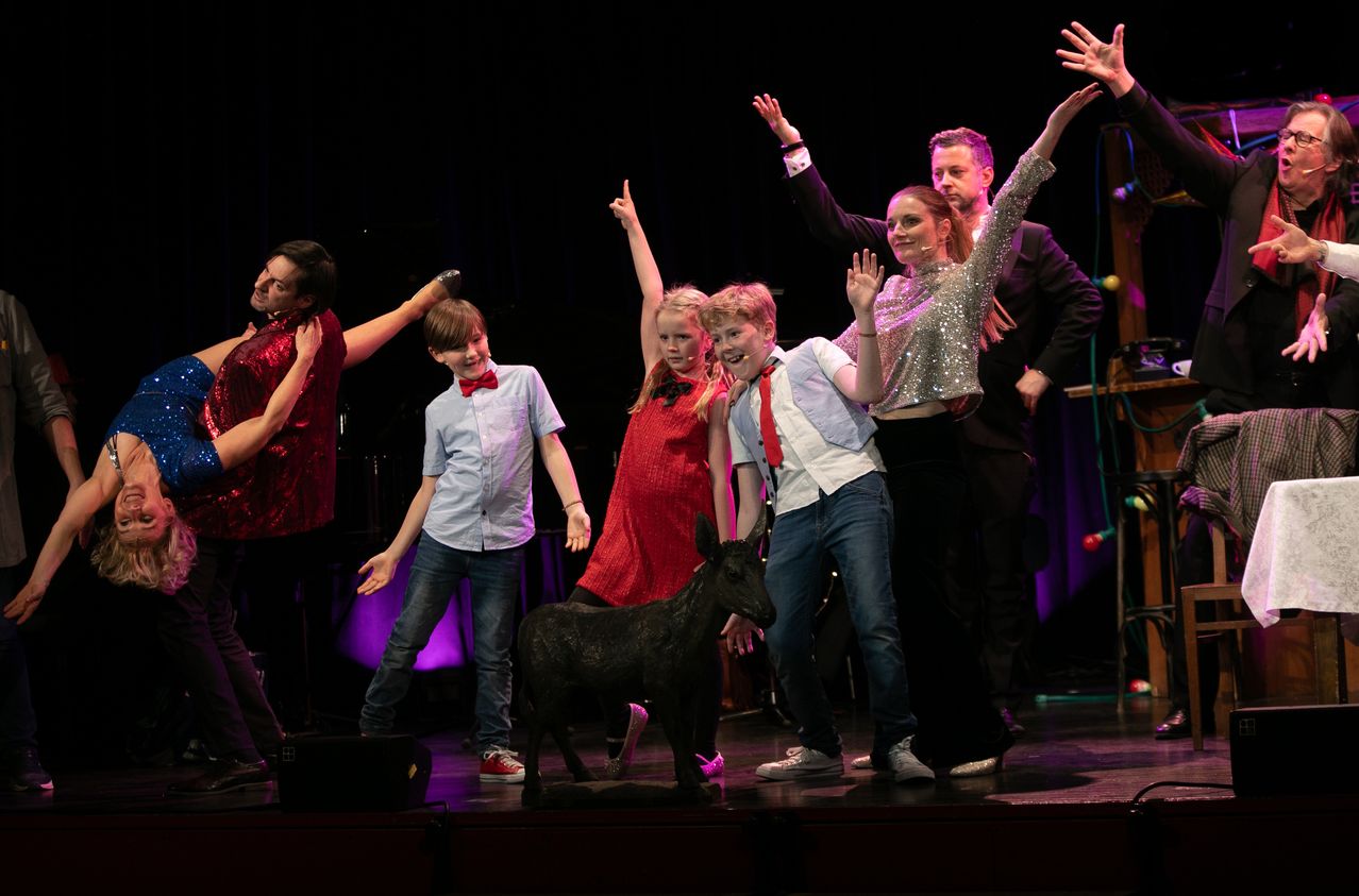 Eine Gruppe von Darstellern steht mit einer schwarzen Ziegenstatue auf der Bühne, trägt festliche Kleidung und winkt. Sie scheinen an einer lebhaften Show teilzunehmen.