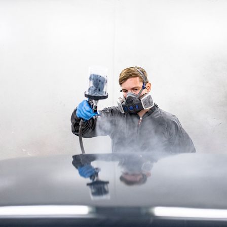 Ein Mann in einem grauen Schutzanzug und Maske sprüht ein Auto mit einem Farbsprüher. Das Auto ist schwarz, und der Mann steht darauf.