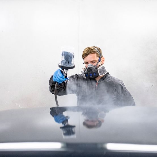 Ein Mann in einem grauen Schutzanzug und Maske sprüht ein Auto mit einem Farbsprüher. Das Auto ist schwarz, und der Mann steht darauf.